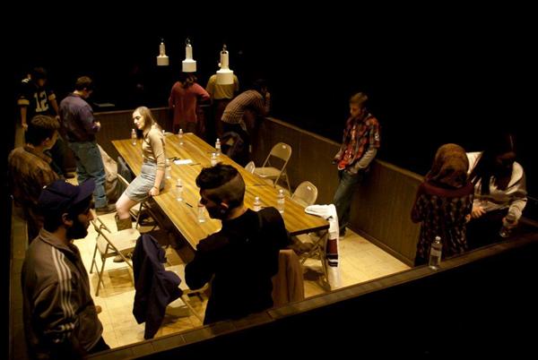 A group of people gather around a long wooden table in a dimly lit room. Some are seated, while others stand and engage in conversation. The setting is intimate, with warm lighting and a few bottles and papers on the table.