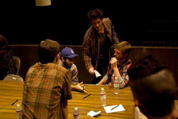 Five men sit around a wooden table scattered with water bottles and pens. One man stands, holding a piece of paper, engaging with the others. The atmosphere is casual, and they appear to be in conversation or discussion.