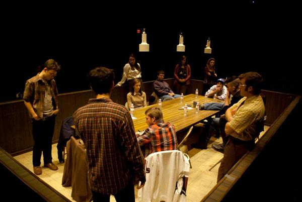A group of people in a dimly lit room gather around a rectangular table. Some are sitting while others stand, engaging in conversation. Hanging lights illuminate the scene, creating a contemplative atmosphere. The setting suggests a meeting or discussion.