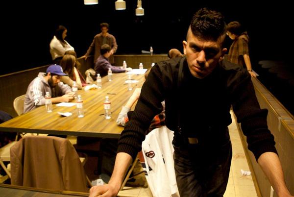 A man leans on a long table in a dimly lit room, with several people sitting and talking around the table. Bottles and papers are scattered on the table, and overhead lights illuminate the scene. The setting appears to be casual or informal.