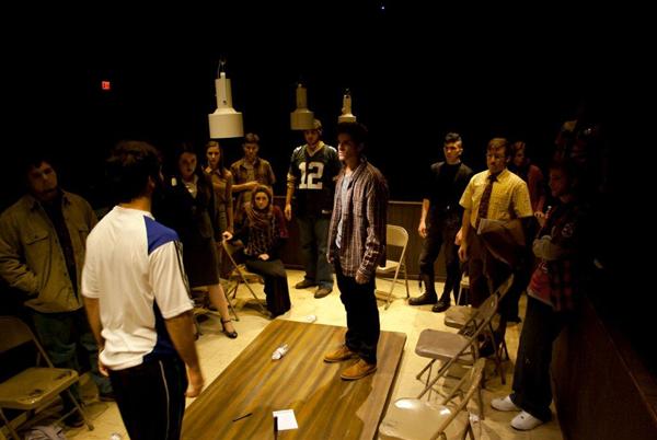 A group of people are gathered in a dimly lit room, standing and sitting in chairs around a table. They appear to be in a serious discussion or confrontation. Some individuals are dressed casually, while others wear more formal attire.