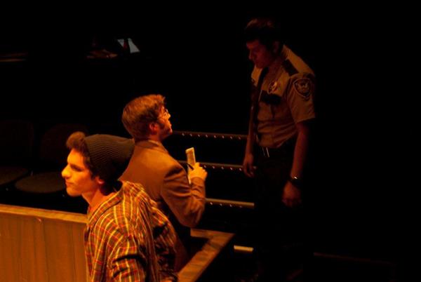 A dimly lit scene with three men. One in a beanie and plaid shirt is in the foreground. Another man in glasses and a suit is seated, holding papers. A third, wearing a brown uniform, stands facing him. The focus is on the seated and uniformed men.