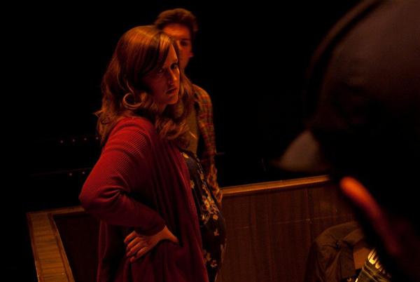 A woman with long hair, wearing a red top, stands with a hand on her hip, looking to the side. She is in a dimly lit room with another person in a plaid shirt partially visible behind her.
