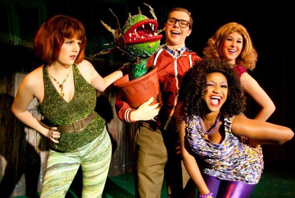 Four people in colorful costumes stand together on stage, smiling. One holds a small potted plant resembling a Venus flytrap with exaggerated features. The group appears to be part of a theatrical performance.