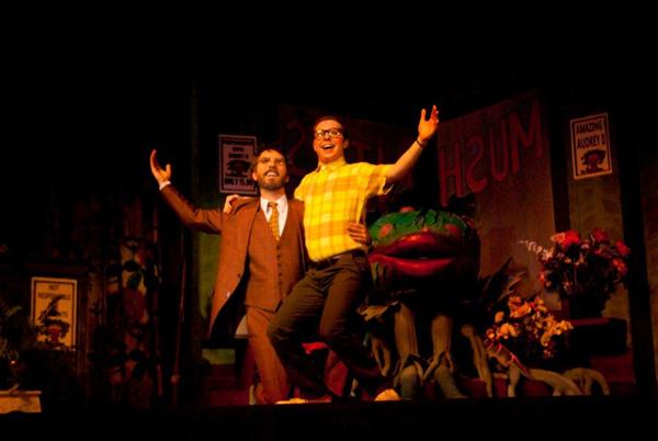 Two actors are on stage, with one lifting the other. One actor is in a suit, and the other wears glasses and a yellow plaid shirt. Behind them is a large plant prop with an open mouth. The setting appears to be from a theatrical production.
