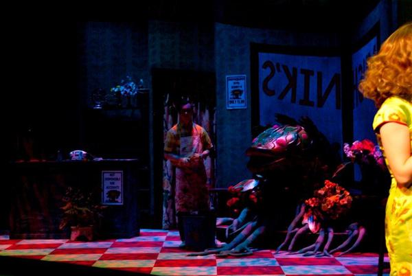 A dimly lit stage scene shows a person in an apron standing next to a large plant with a gaping mouth and tentacle-like roots. Posters on the wall read "Snik." A woman with blonde hair stands on the right. The floor has a checkered pattern.