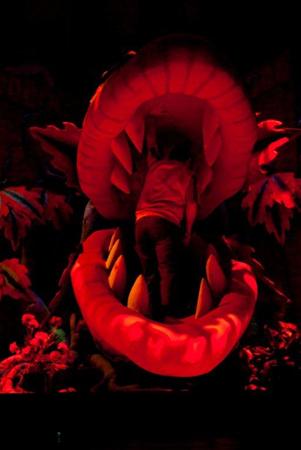 A person appears to be entering a giant, open-mouthed plant with large teeth and red lighting, suggestive of a scene from a theatrical production. Colorful leaves surround the mouth, adding a dramatic, fantasy-like atmosphere.