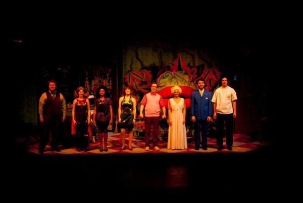 A group of eight people standing on a stage, possibly after a performance. They are dressed in colorful and formal attire, and the stage backdrop includes a large colorful pattern. The lighting is dim, focusing on the individuals.