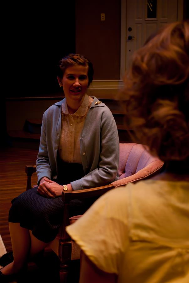 Two women sit in a warmly lit room. One, with short hair, wears a light-colored blouse and blue cardigan, sitting on a pink chair. The other woman, with curly hair, is seen from the back. The setting is cozy and intimate.