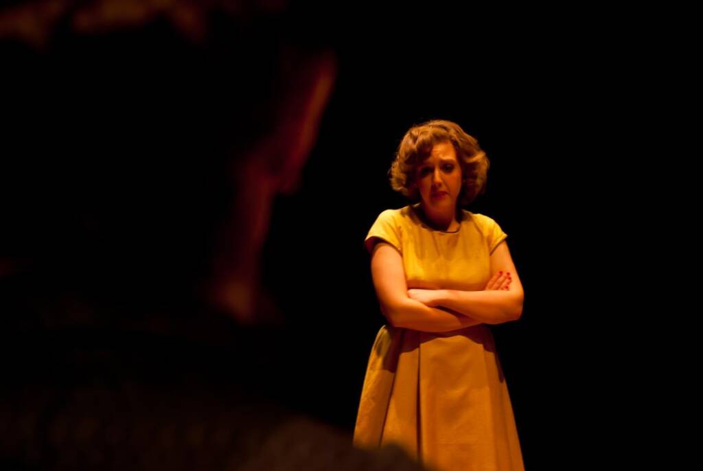 A woman in a yellow dress stands on a dimly lit stage with her arms crossed, looking downward. The background is dark, and the scene has a dramatic, theatrical atmosphere.