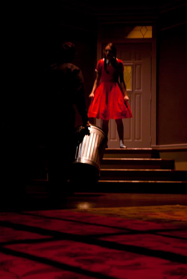 A person in a red dress stands on steps in front of a door, looking towards another person holding a metal trash can in a dimly lit environment.