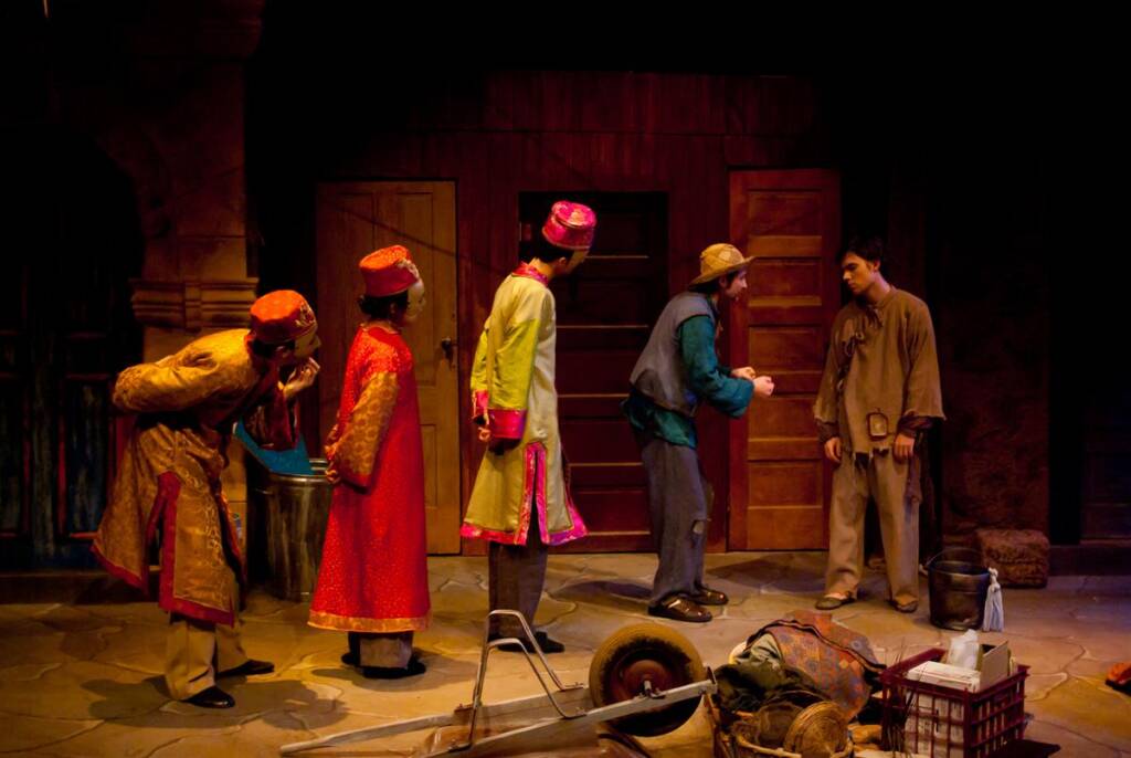 Actors in colorful costumes perform a scene on stage. Four people in traditional attire interact with one person in plain clothing near wooden doors. Props, including a wheelbarrow, are on the floor in front of them.