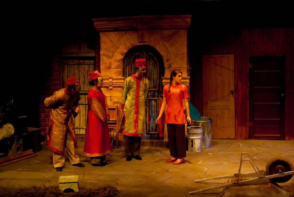 Four actors in colorful traditional costumes perform on stage with a backdrop resembling a rustic street. One actor, dressed in a modern orange outfit, stands facing the others. Various props, like a wheelbarrow and barrels, are scattered around.