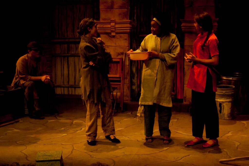 Four actors perform on a dimly lit stage set, resembling an old rustic room. Three stand engaged in conversation, with one holding a bowl. The fourth actor sits on the left, watching. The scene conveys a dramatic and contemplative mood.