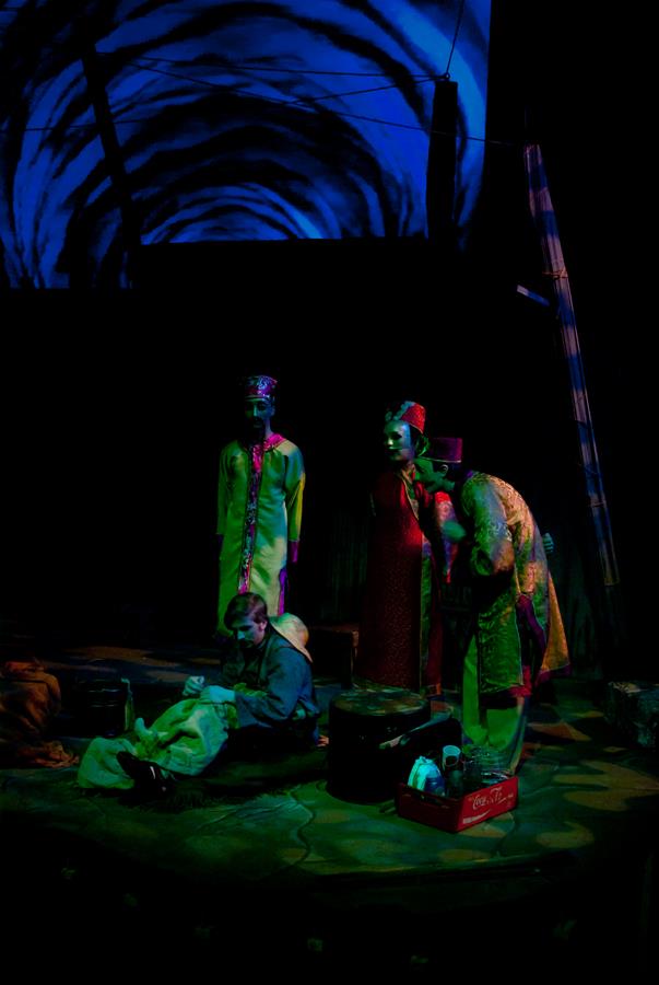 A dimly lit stage shows a scene with theatrical performers in colorful costumes gathered around a man seated on the ground. One performer leans in, appearing concerned. The background features blue swirling patterns. Props include a crate and a small box.