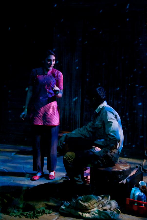 A woman in a red dress stands and gestures toward a seated man in a dimly lit room. Light blue spots resemble snowflakes around them. The man sits on a stool beside scattered clothing and objects on the floor.