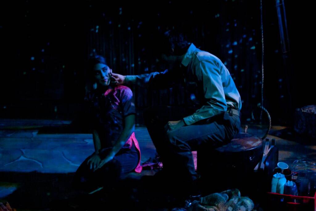 A dimly lit scene with two people on a stage. One person is sitting on the ground, wearing a pink shirt and scarf. The other, seated on a bucket, gently touches the person's face. Various props surround them, with a blue and pink light creating a dramatic effect.