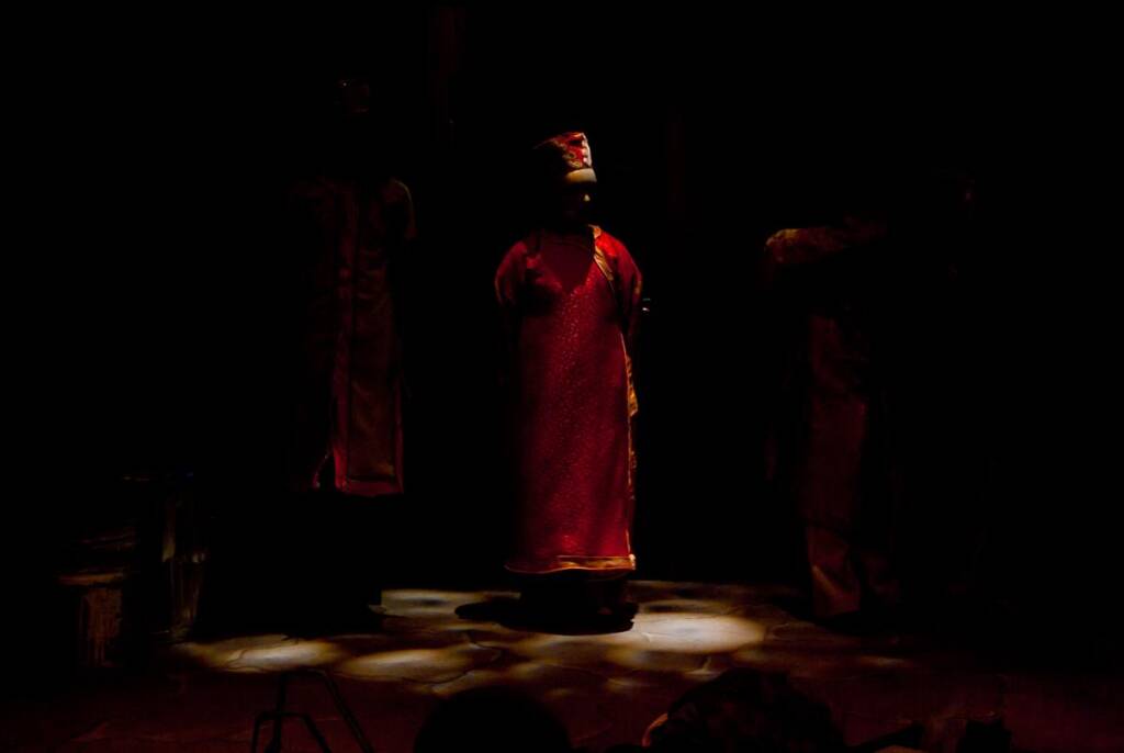 A person stands in the center of a dimly lit stage, wearing a long, red traditional outfit with a matching hat. The person is illuminated by a spotlight, while two other shadowy figures are partially visible in the background.