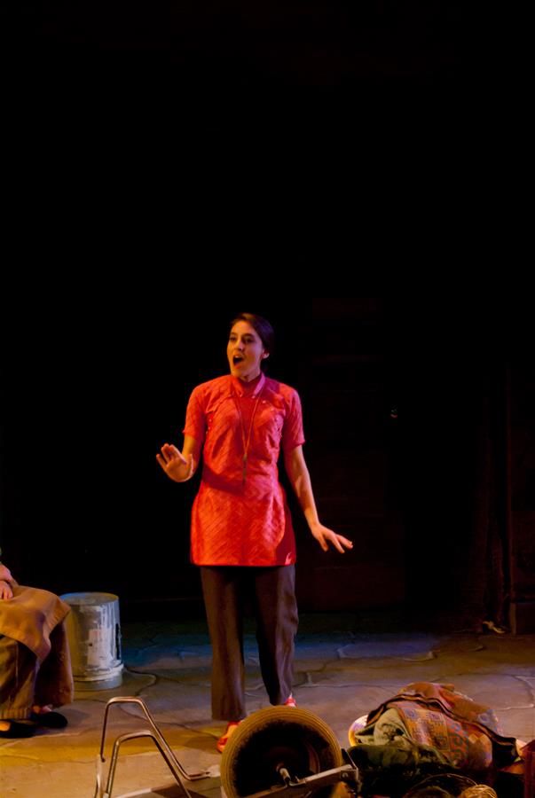 A woman in a red top and dark pants stands on a dimly lit stage, gesturing with one hand. A metal bucket and some scattered objects are on the floor near her. She appears to be performing or speaking.