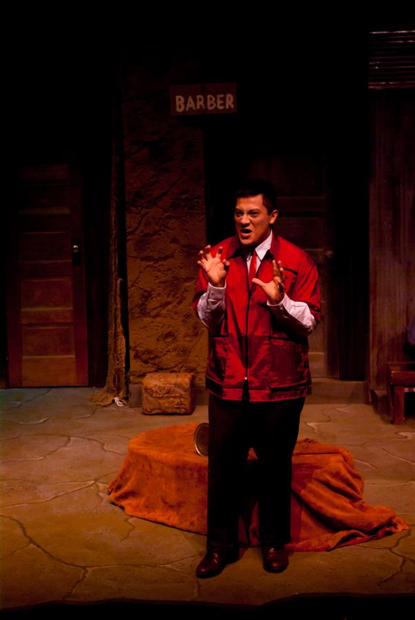 A person wearing a red jacket performs on stage, set against a rustic backdrop with a "Barber" sign. The lighting is dim, creating a dramatic ambiance. The performer gestures expressively, engaging with the audience.