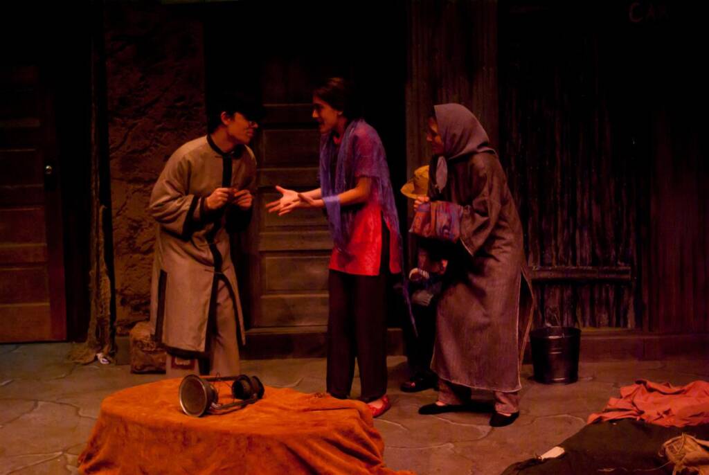 Three actors perform on a dimly lit stage. One in a long coat and hat gestures animatedly, another in a red top and slacks responds with open hands, and an older person with a shawl stands nearby, holding a wrapped bundle.
