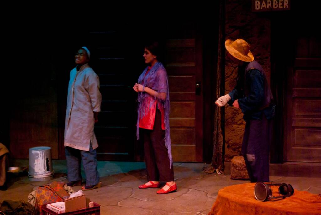 Three actors on a dimly lit stage perform a scene. One wears a long coat and hat, another is in a shawl with red shoes, and the third wears a wide-brimmed hat. A "BARBER" sign and props like a metal bucket and books are visible.