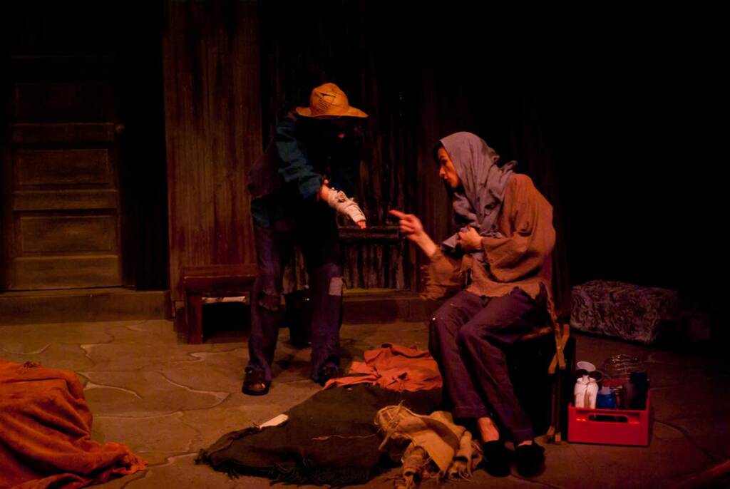 Two actors perform on a dimly lit stage. One, dressed in a hat and coat, points at the other, who sits in shabby clothing and a headscarf. Props and blankets are scattered around, creating a rustic setting.