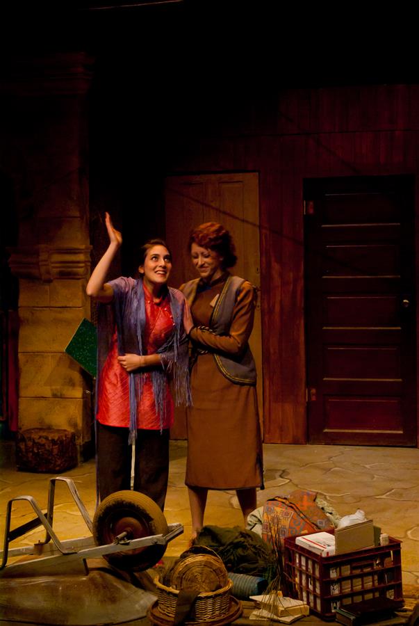 Two women stand on a stage, one gesturing upwards with a smile. They are surrounded by a disorganized pile of props including a wheelbarrow and a hat. The background features a partially open door and wooden paneling.