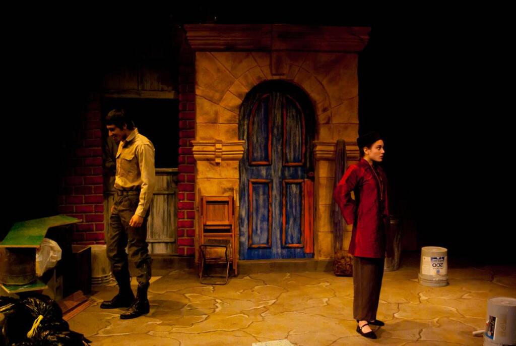 A dimly lit stage scene with two actors. A man in a yellow shirt and green pants looks down near scattered props. A woman in a red jacket stands with hands behind her back, facing away. A rustic backdrop features a blue door and brick walls.