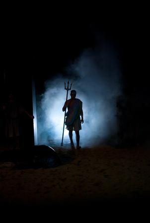 A person in a toga-style outfit holds a trident, silhouetted against a misty blue light in a dark setting. The scene evokes a dramatic or mythical atmosphere.