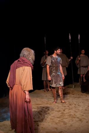 A theatrical scene with actors in ancient costume. A person in a long robe stands in the foreground, facing another wearing armor and sandals. In the background, three individuals hold spears, dressed in tunics. The stage floor appears sandy.