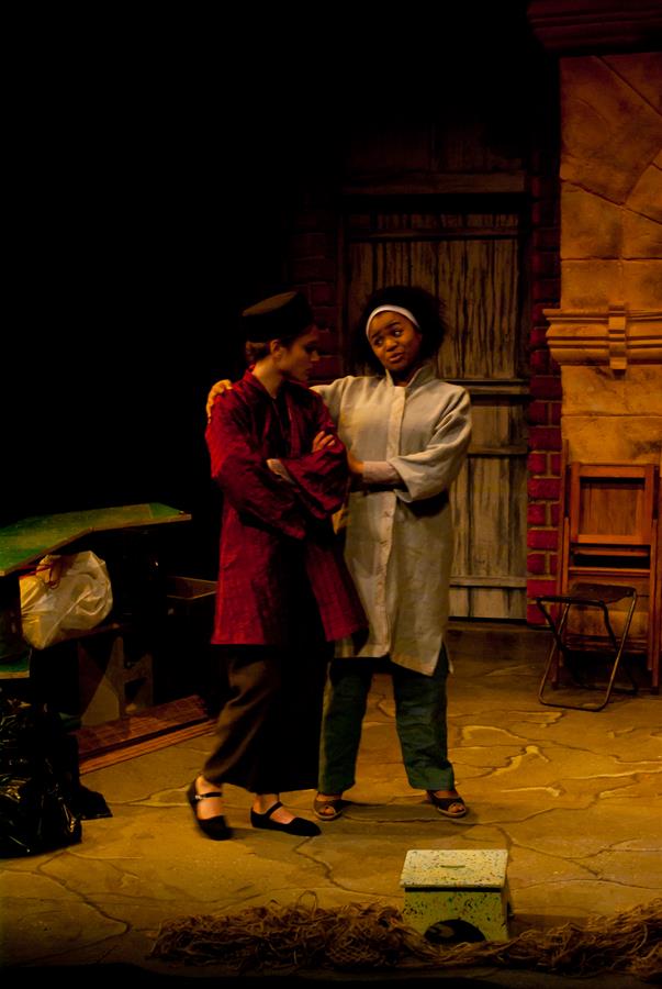 Two actors perform on a dimly lit stage. One wears a red robe and black hat, with crossed arms; the other wears a light coat and black hat, gesturing with one arm around the first actor's shoulder. Behind them is a textured wall with a door.