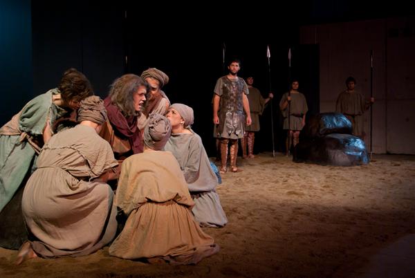 A theatrical scene with actors in desert-themed costumes. One person sits on the ground surrounded by others, appearing distressed, while a standing figure observes. The stage is sandy with props resembling rocks and spears in the background.