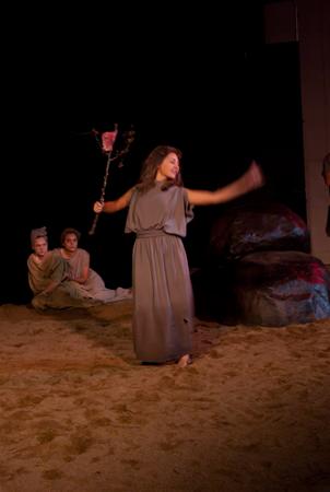 A person in a long dress holds a staff while performing on a dimly lit stage with a sandy floor. Three seated people watch from the background near large rocks. The atmosphere suggests a theatrical or dramatic scene.