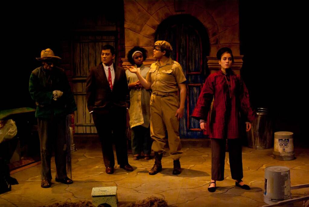 Five performers stand onstage portraying different characters. One wears a hat and glasses, another a suit, two in casual costumes, and one in a uniform with a cap. The set resembles a rustic, dimly lit building, enhancing the dramatic atmosphere.