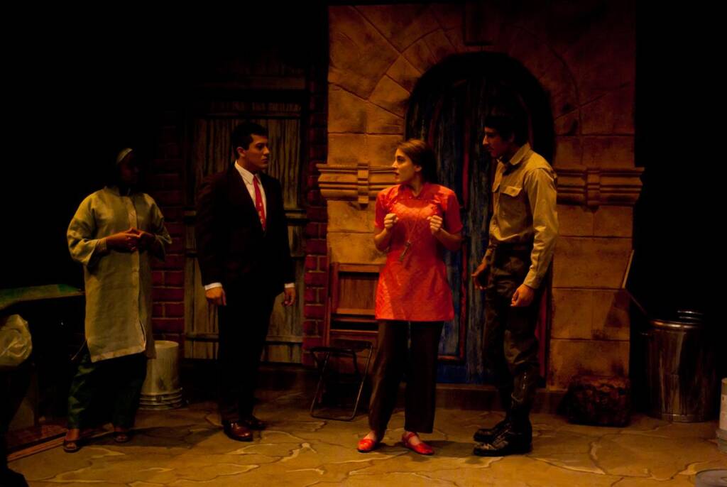 A theatrical scene with four actors in costume on stage. The set is designed to resemble an alley with doors and a stone wall. One actor in a dress gestures animatedly, while the others wear a suit, uniform, and apron, observing her.
