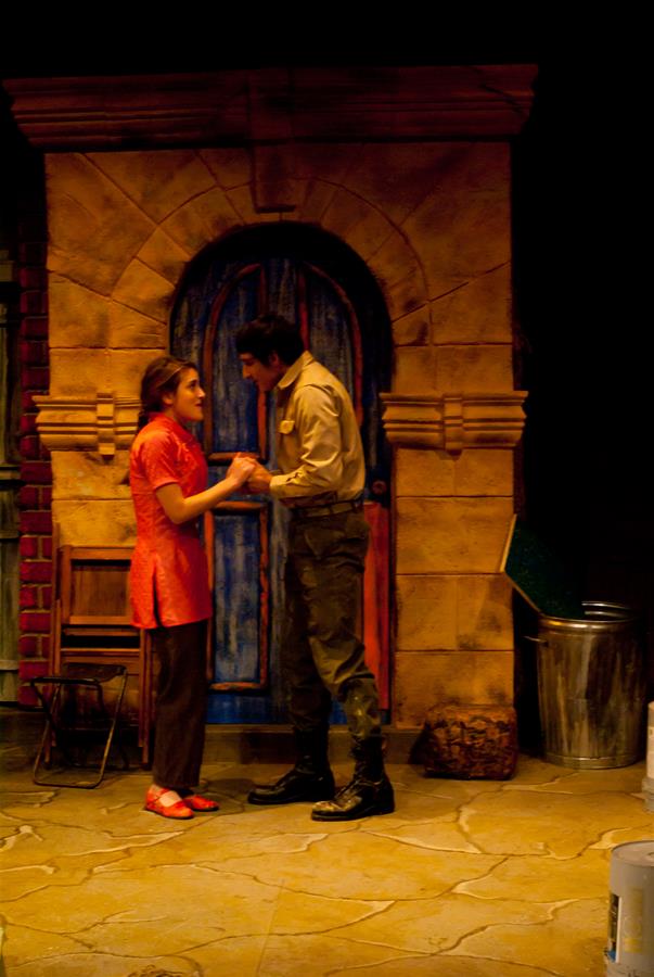 A woman in a red dress and red shoes holds hands with a man in a brown uniform in front of an arched, blue door. They are on a dimly lit stage with props including a trash can and a wooden chair.