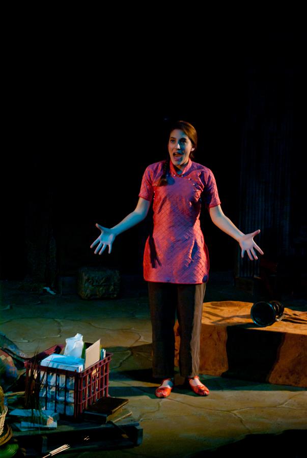 A woman stands on a dimly lit stage, wearing a pink tunic and pants. Her arms are outstretched, and she appears to be speaking or singing. A crate with items is visible on the stage floor.