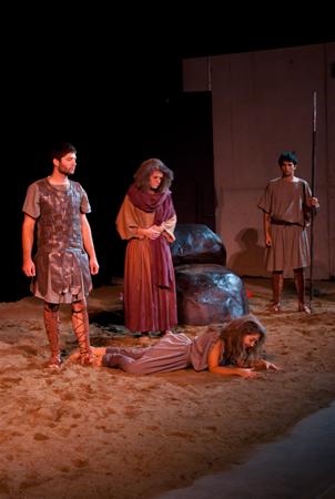 A theatrical scene on stage features four actors in ancient-style clothing. One actor lies on the ground, while the other three stand nearby. The backdrop includes large rocks and a partial wall. The lighting casts dramatic shadows.