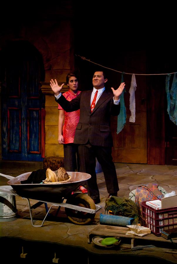 A man in a suit gestures dramatically on stage, flanked by a woman in a red dress. A wheelbarrow filled with various items sits nearby. The background features a rustic setting with hanging laundry and a closed door.