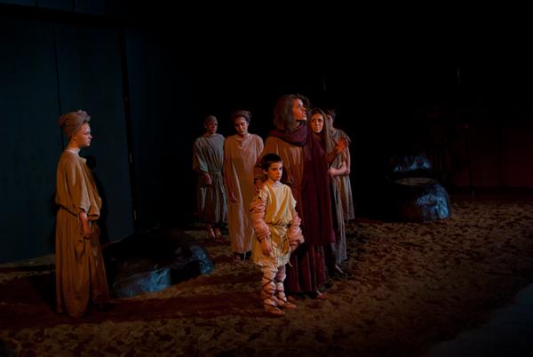 A group of people in historical or biblical costumes stand on a dimly lit stage resembling a sandy desert. The scene includes children and adults, with one central figure surrounded by others, evoking a dramatic or theatrical atmosphere.