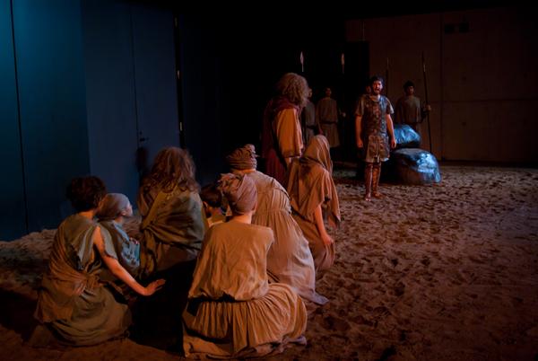 A group of people in brown clothing kneel on a sandy stage, facing a standing man in a tunic and armor. Dim lighting suggests a dramatic theater setting with others in the background.