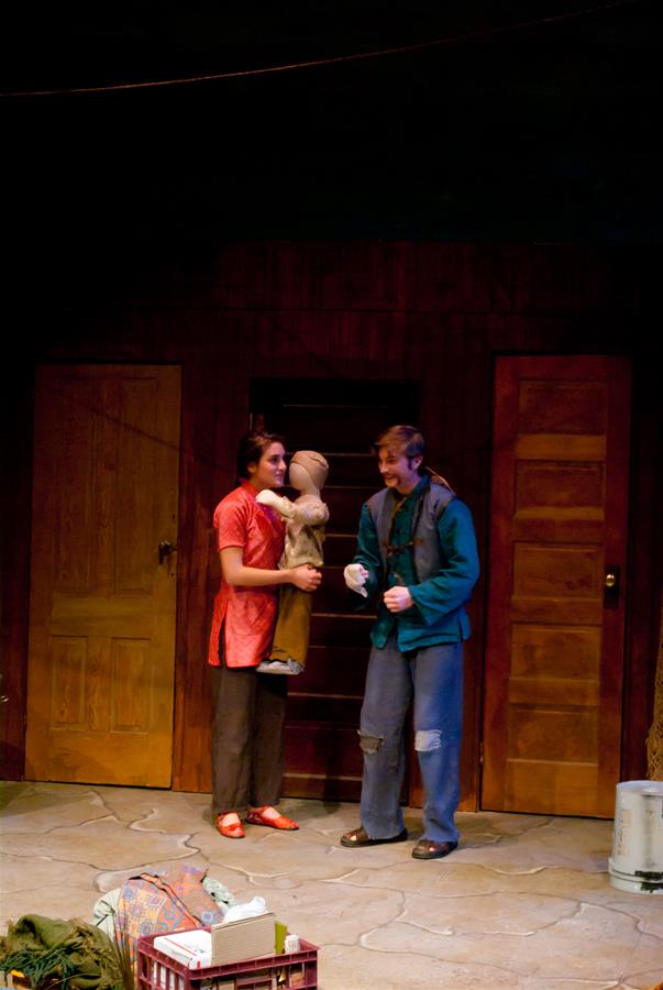 Two performers are on stage; one holds a puppet resembling a child. The background features wooden doors. The scene is set on a stone-patterned floor with a basket of props in the foreground.