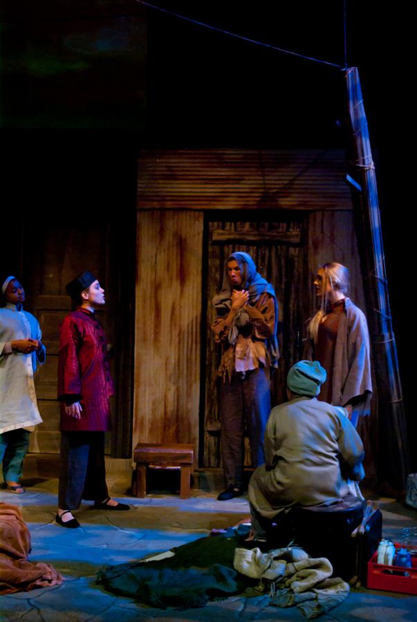 A theatrical stage scene with five actors. One actor sits on the floor, while two stand facing each other in conversation. The setting includes a rustic-looking backdrop and scattered props, creating a dramatic atmosphere.