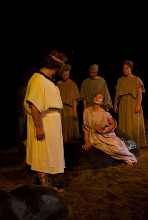 A theatrical scene features five people in ancient Greek-style costumes. One person sits on a rock, looking up at another person who is standing, while the others watch. The dim lighting gives a dramatic, historical ambiance.
