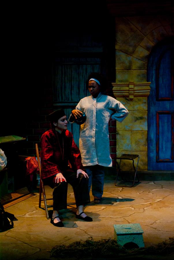 Two performers are on stage. One, seated, wears a red and black outfit and a hat. The other stands, gesturing, dressed in a white coat. They are set against a backdrop of a doorway and brick wall. The lighting is dim, focusing on the actors.