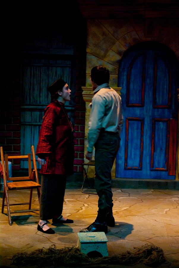 Two actors are in a dramatic scene on stage. One wears a red jacket and beret, and the other in a beige shirt and boots. They are standing near a wooden chair and a blue, arched door in a dimly lit setting.
