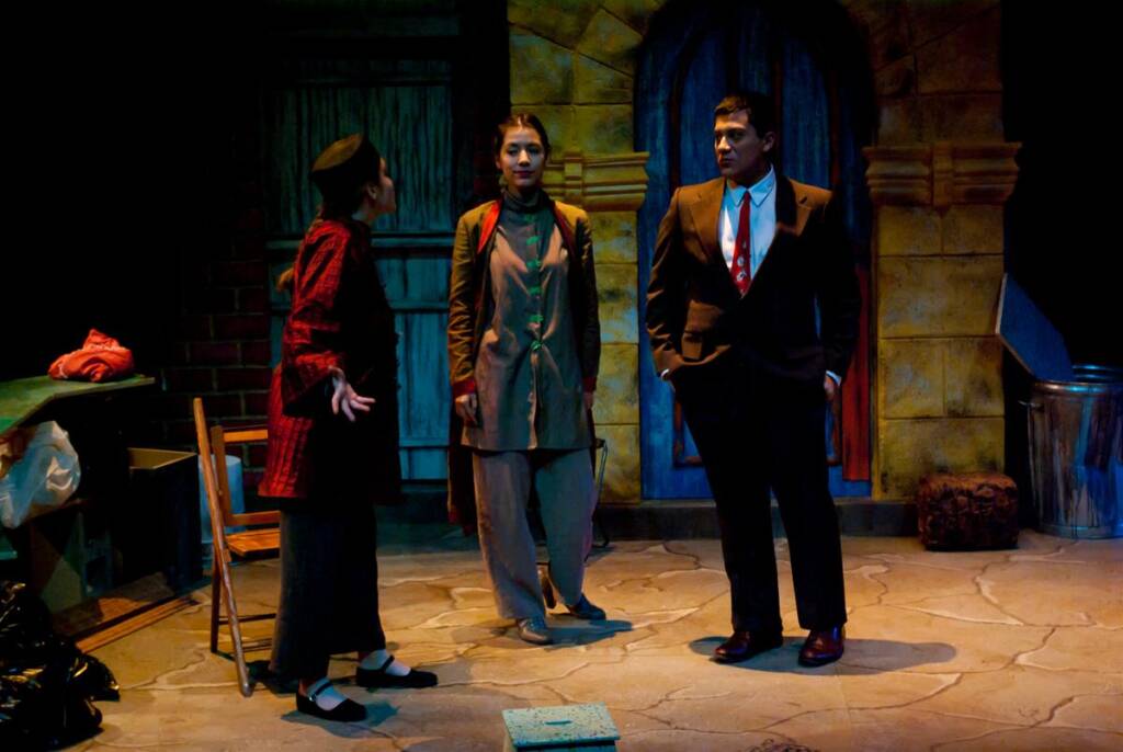 Three people are performing on stage. One, a woman in a red plaid shirt, gestures passionately. The others, dressed in brown and a suit, look on. The setting resembles an old street with arched doors and stacked props.