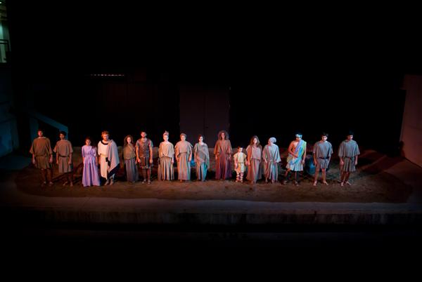 A group of people dressed in simple, earth-toned costumes standing on a stage. They appear to be participating in a theatrical performance, lined up and facing the audience under dim lighting.