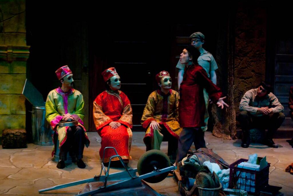 A theatrical scene with four actors in colorful robes and masks seated, while another actor in red stands gesturing dramatically. Two people in plain clothes, one standing and one sitting, watch them. A wheelbarrow and scattered props are in the foreground.